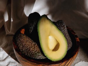 Primer Plano De Un Aguacate Hass Cortado Por La Mitad, Mostrando Su Pulpa Cremosa En Un Tazón De Madera, Con Otros Aguacates En Segundo Plano. Fotografía De Aguacates Frescos, Ideal Para Aguacates A Domicilio En Barcelona Y Madrid.